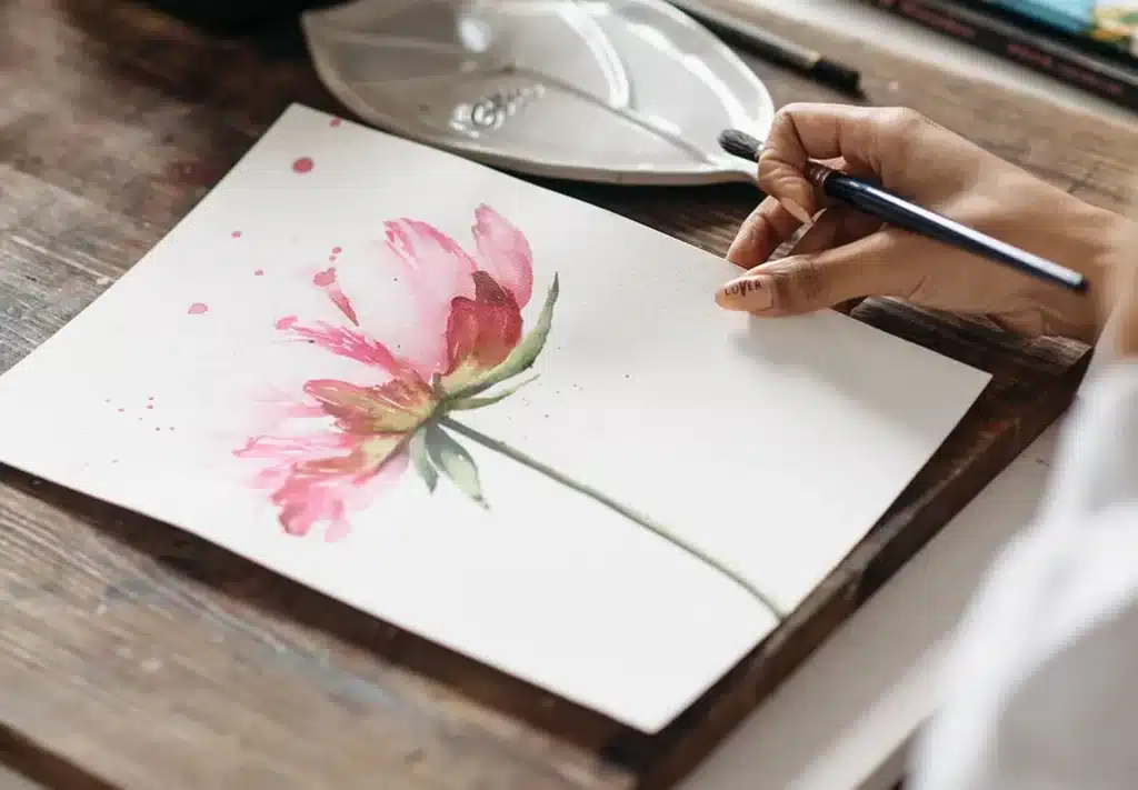 A hand holding a paintbrush drawing a watercolor flower on a white paper