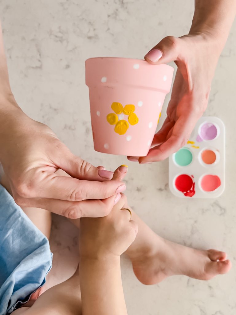 A person holding the finger of a child with paint on it while painting a pot plant