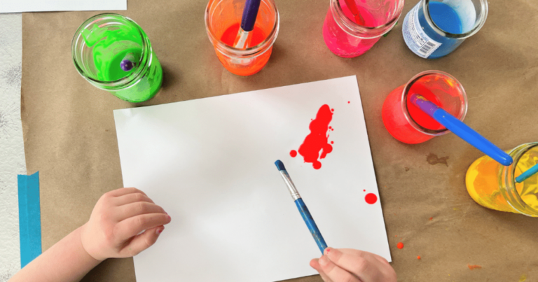 A top view of child's hands holding a paint brush over a white paper with red paint with colored water all around