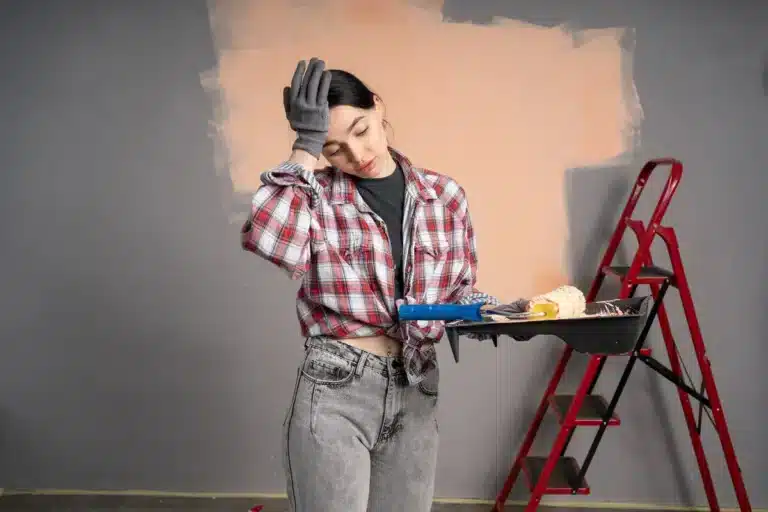 a girl seems worried holding a paint roller a half painted wall on back