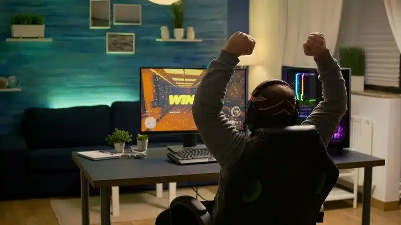 A gamer celebrating a victory in front of their setup with a colorful background and modern gaming equipment