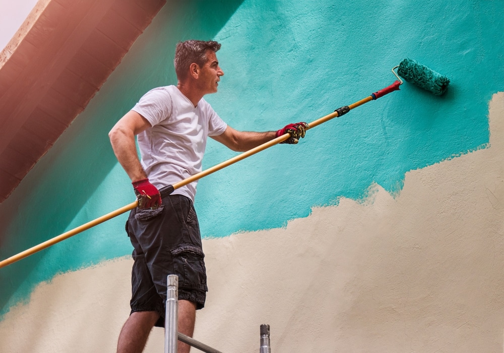 A view of a person painting an exterior wall with a roller A view of a person painting an exterior wall with a roller
