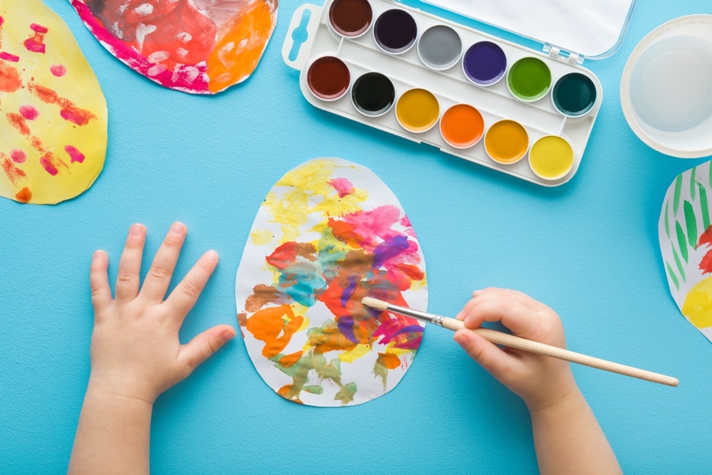 Baby Boy Holding Paintbrush And Painting Colorful Egg On Paper Baby Boy Holding Paintbrush And Painting Colorful Egg On Paper